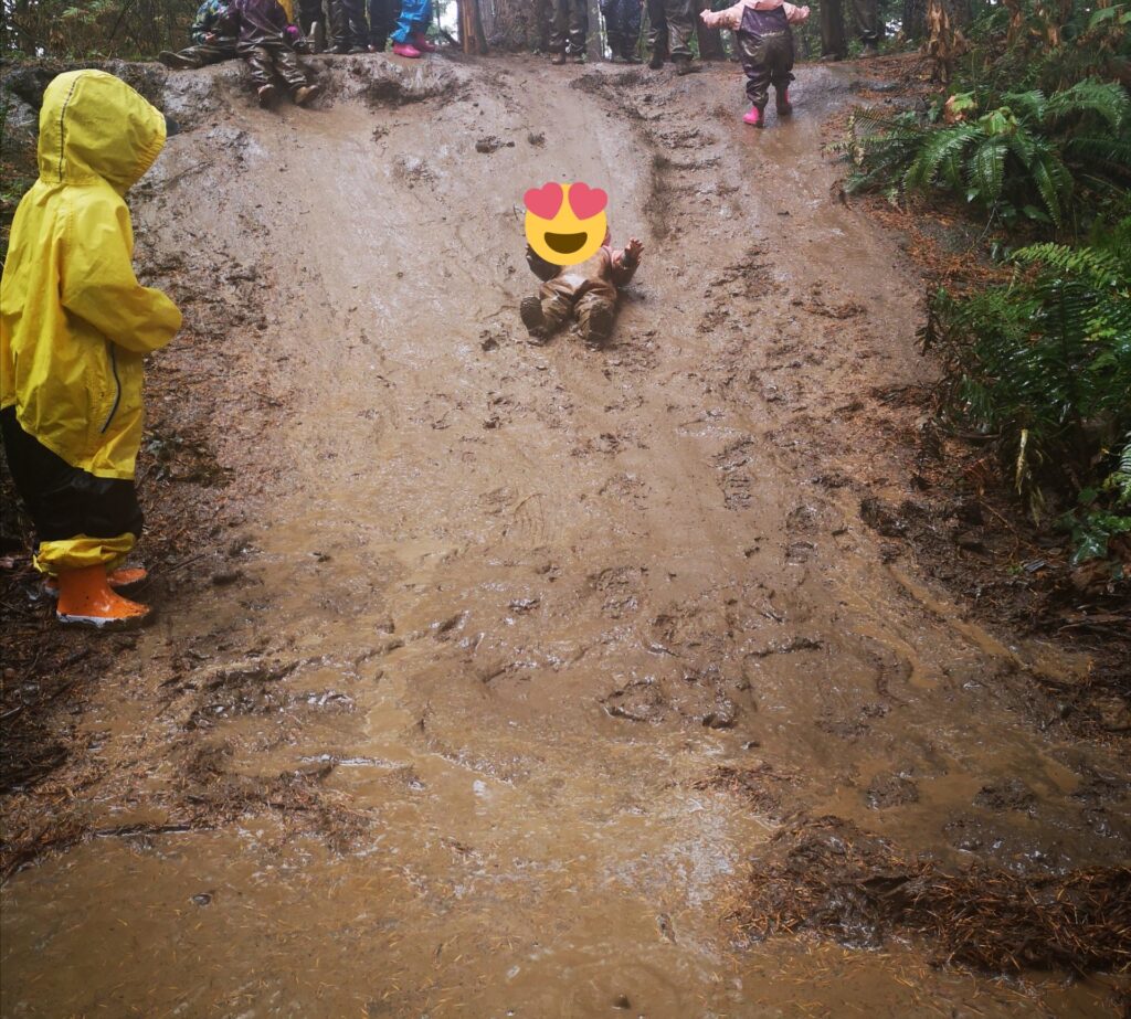 雨の中森の中に自然にできた泥の滑り台を子どもが滑るようす